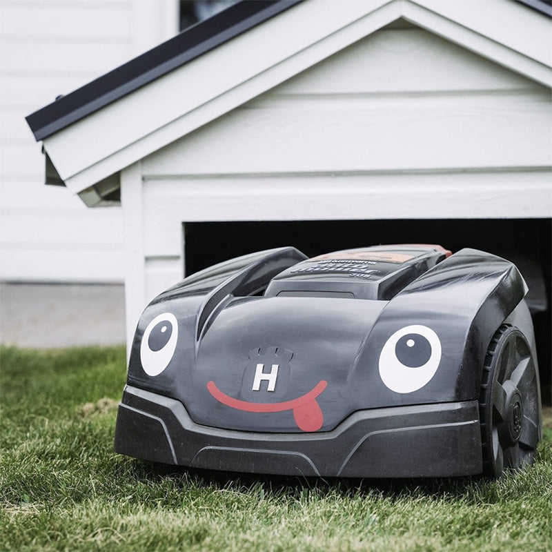 Robotgräsklippare med ögon och mun framför garage
